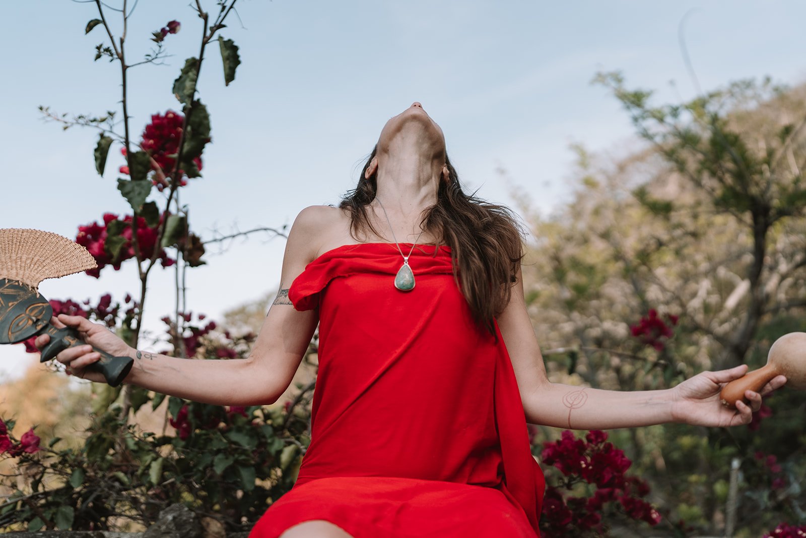 A woman in a red dress with a green stone pendant is outdoors among plants and flowers, holding a fan in one hand and an object in the other, with her head tilted back and eyes closed.