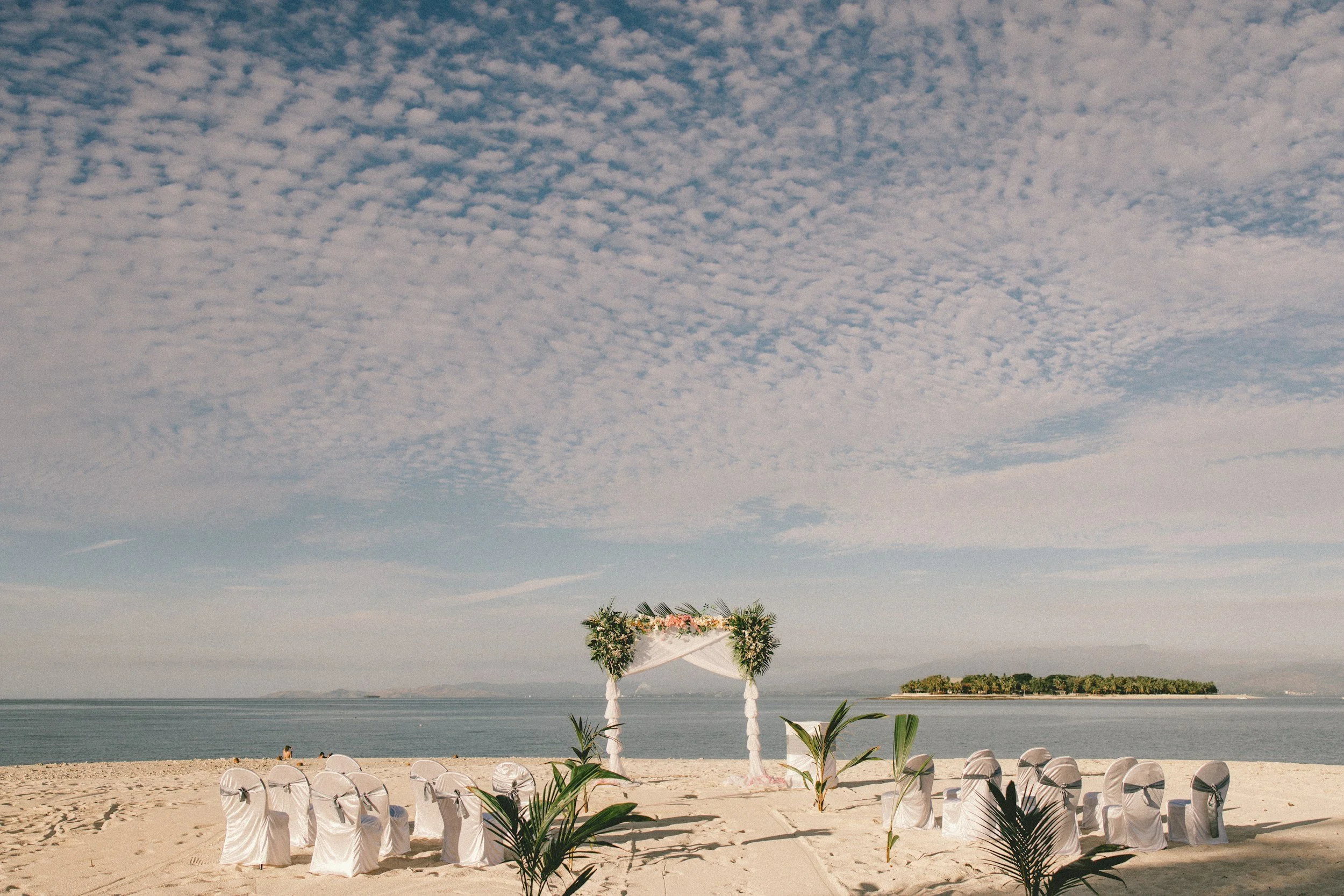 Beach wedding setup with white chairs, a floral arch, and tropical plants under a cloudy sky with an island in the background.