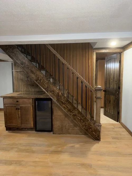 Wooden staircase next to a wooden cabinet and a mini-fridge in a room with wood flooring and paneling.