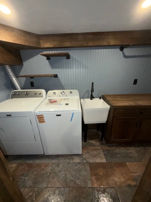 Laundry room with a washing machine and a dryer next to a utility sink, dark wood countertop, floating shelves on the wall, and tiled floor.