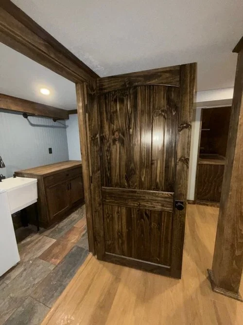 A wooden door partially opened in a room with wood and stone flooring, and wooden paneling, revealing a small closet space.