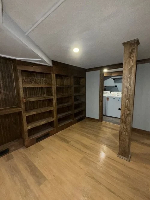 Empty room with wooden floors, built-in wooden bookshelves, and a laundry area with washing machines in the background.