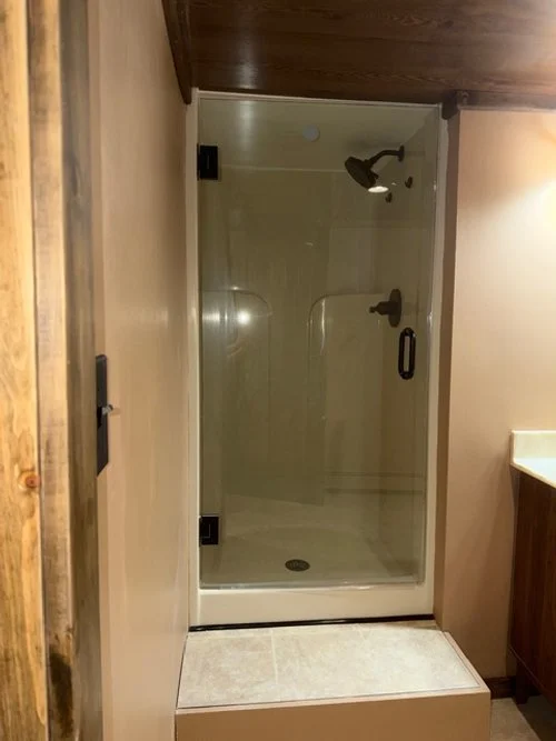 Glass shower enclosure with a showerhead and handle, surrounded by beige tiles and wooden fixtures.