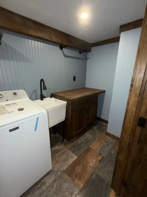 Laundry room with a washing machine, a utility sink, and a wooden cabinet on tiled flooring and a wall with vertical beadboard paneling.