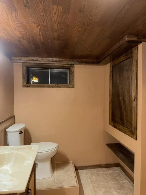 Small bathroom with a wooden ceiling, beige walls, and a small window. It has a white toilet, a beige sink, and a dark wood shelf and frame on the wall.