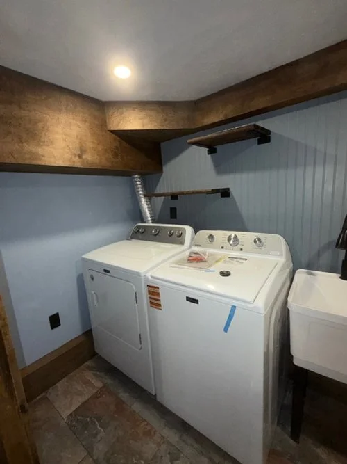 A laundry room with a washer and dryer, painted blue walls, two wooden shelves, a beige ceiling with a recessed light, and a small utility sink.