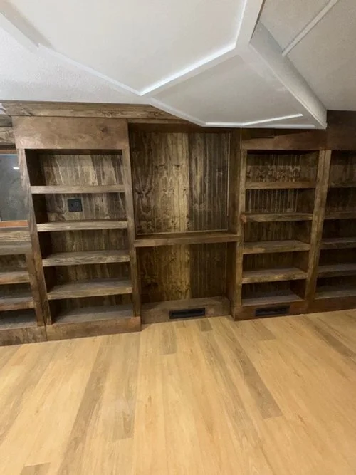 Empty wooden built-in bookshelves with five shelves each, set against a dark wood-paneled wall in a room with light wooden flooring.