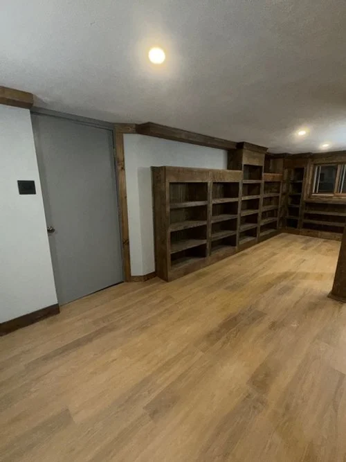Empty room with built-in wooden bookshelves along the wall, hardwood floor, white walls, gray door, and ceiling lights.