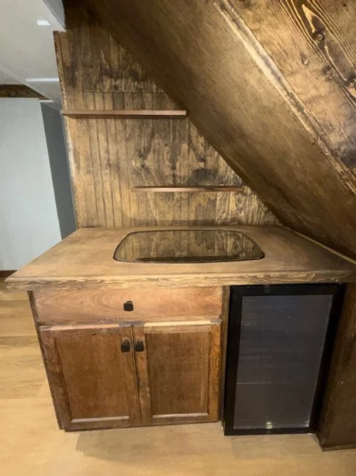 A small wooden kitchenette with a countertop, a built-in stovetop, and a mini fridge under the counter, installed under an angled wooden ceiling.