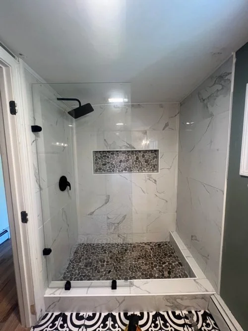 Custom walk-in shower remodel with marble tiled walls, pebble stone floor, built-in shower niche, and a glass door enclosure installed by P Jordan Construction in New Hampshire