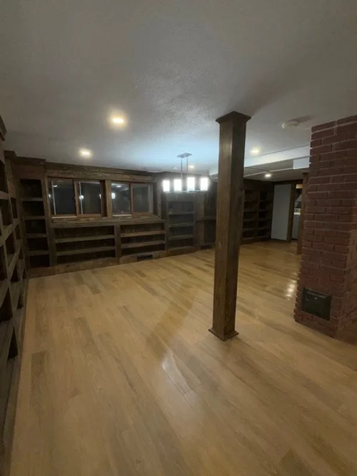 Empty room with wooden floors, built-in wooden shelves along two walls, a brick chimney or wall with a vent, a wooden support beam, and a modern ceiling light fixture.