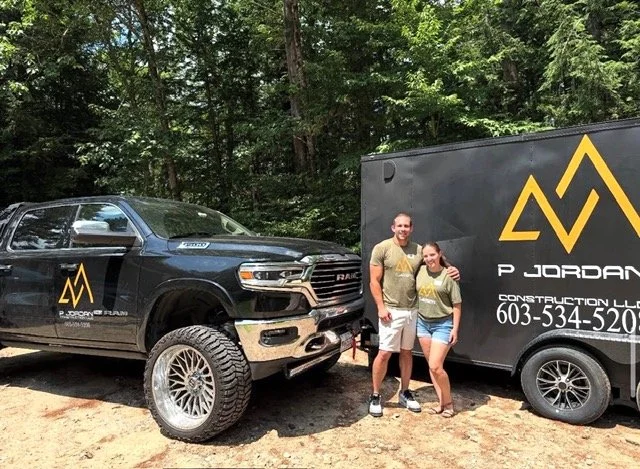A man and woman standing next to a black pickup truck and a black construction trailer, both with yellow mountain logo and text, in a wooded outdoor setting.