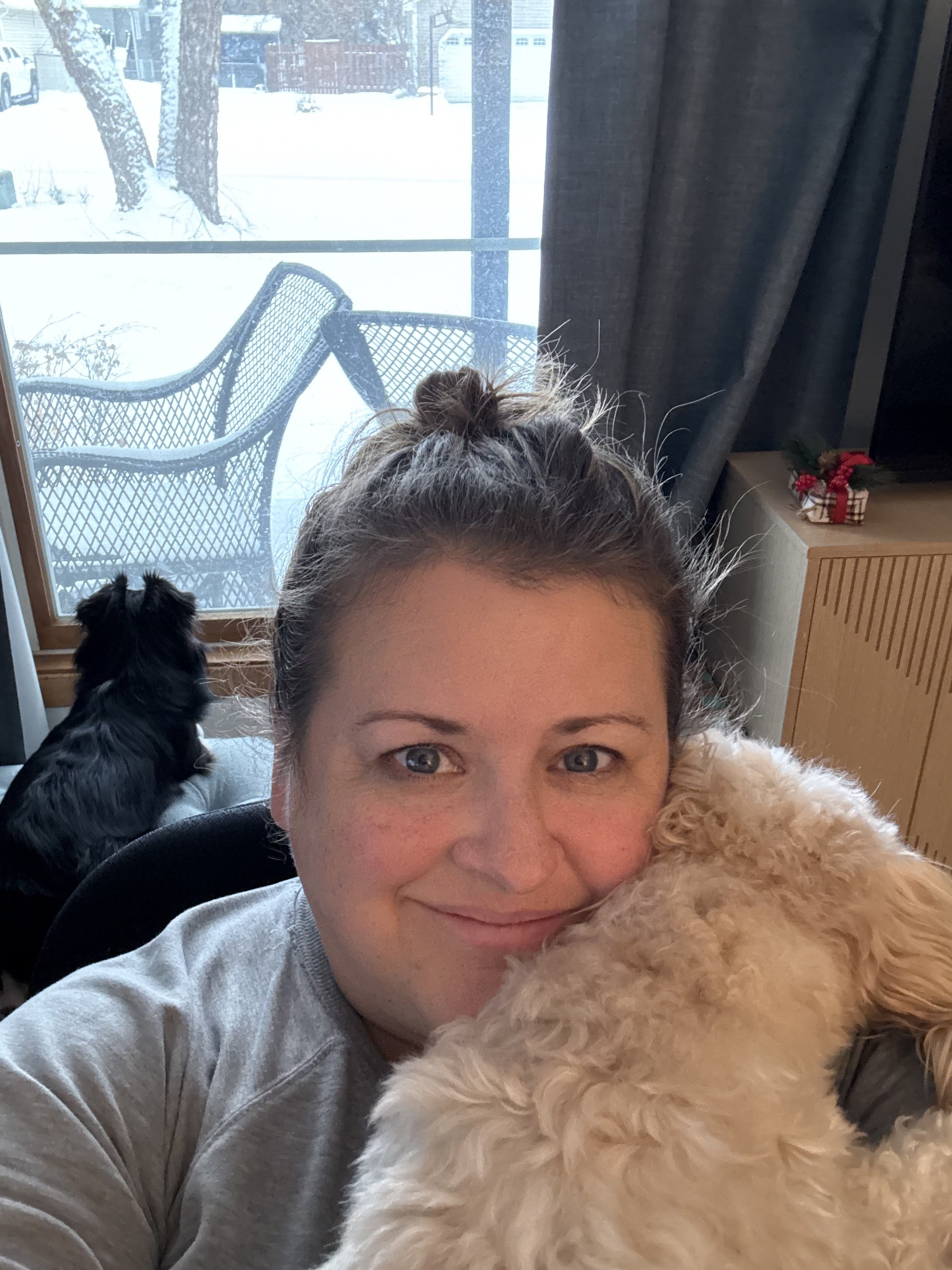 A woman with gray hair in a ponytail and a young woman with curly hair holding a light-colored fluffy dog close to her face inside a home, with a snow-covered backyard and patio furniture visible through a sliding glass door in the background.