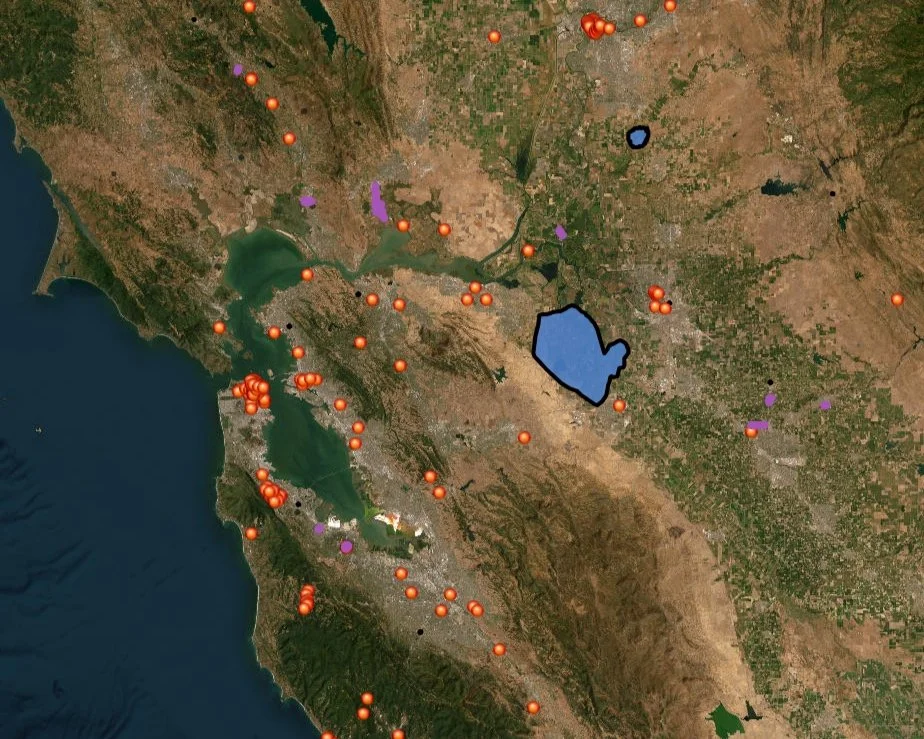 Map showing various points of interest with orange markers around a large body of water, possibly a reservoir or lake, with surrounding terrain and smaller water bodies. There are some purple and green markers, and the background features a mix of urban and rural areas.