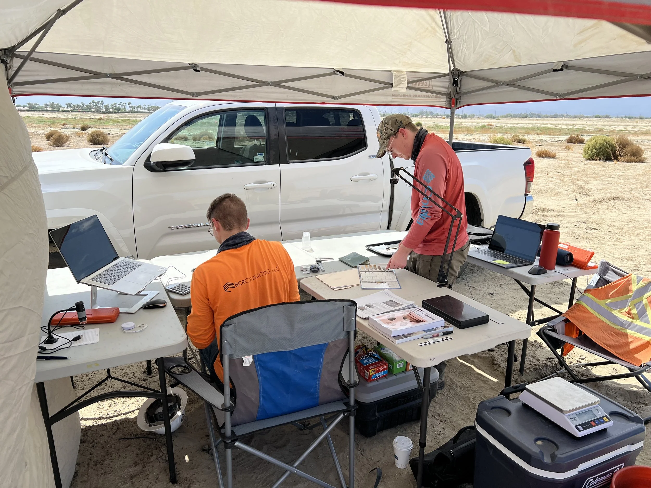 Two BCR team members under a canopy tent outdoors with a white pickup truck in the background, working at tables with laptops, documents, and equipment in a dry, desert-like area.