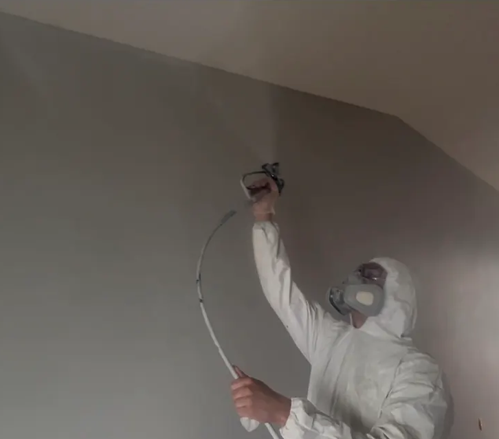 A person in full protective gear, including a mask and hazmat suit, using a spray gun to disinfect or treat a ceiling.