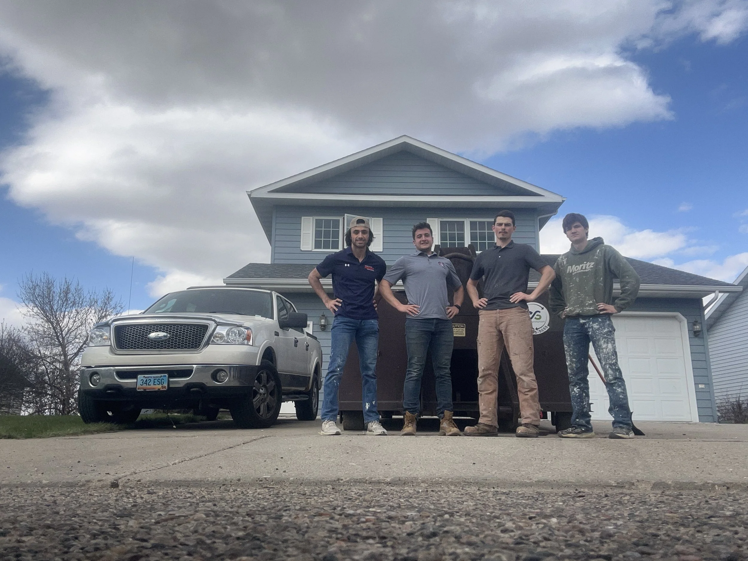 Four young men standing side by side in front of a blue two-story house with white shutters, a garage, and a driveway. A white truck is parked in front of the house, and a black trash bin is visible behind them. The sky is cloudy with some patches of blue.