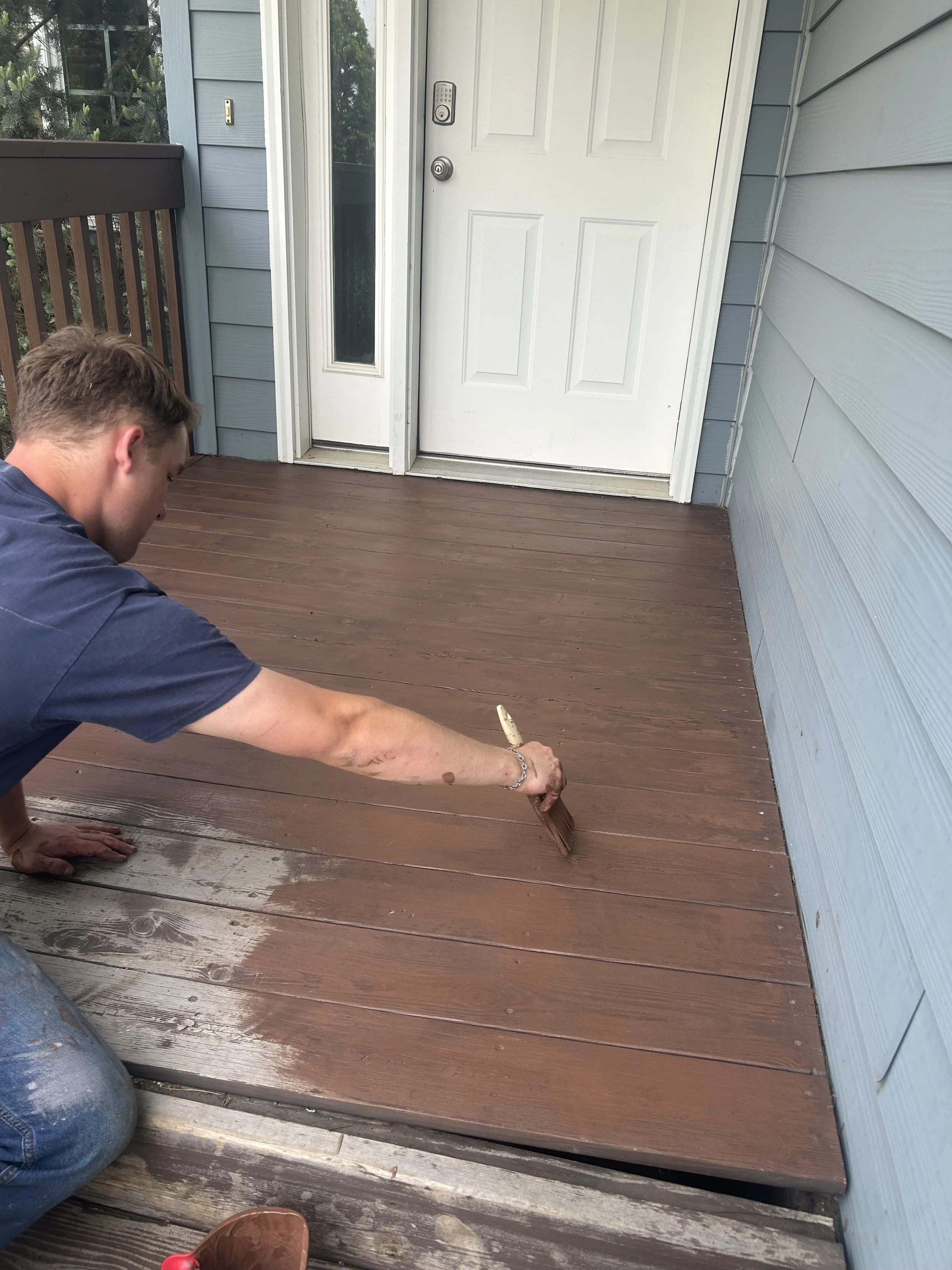 A person with short brown hair is kneeling on a wooden porch, applying a brown stain to the deck with a brush. The porch is attached to a house with light blue siding and a white door with a vertical window.