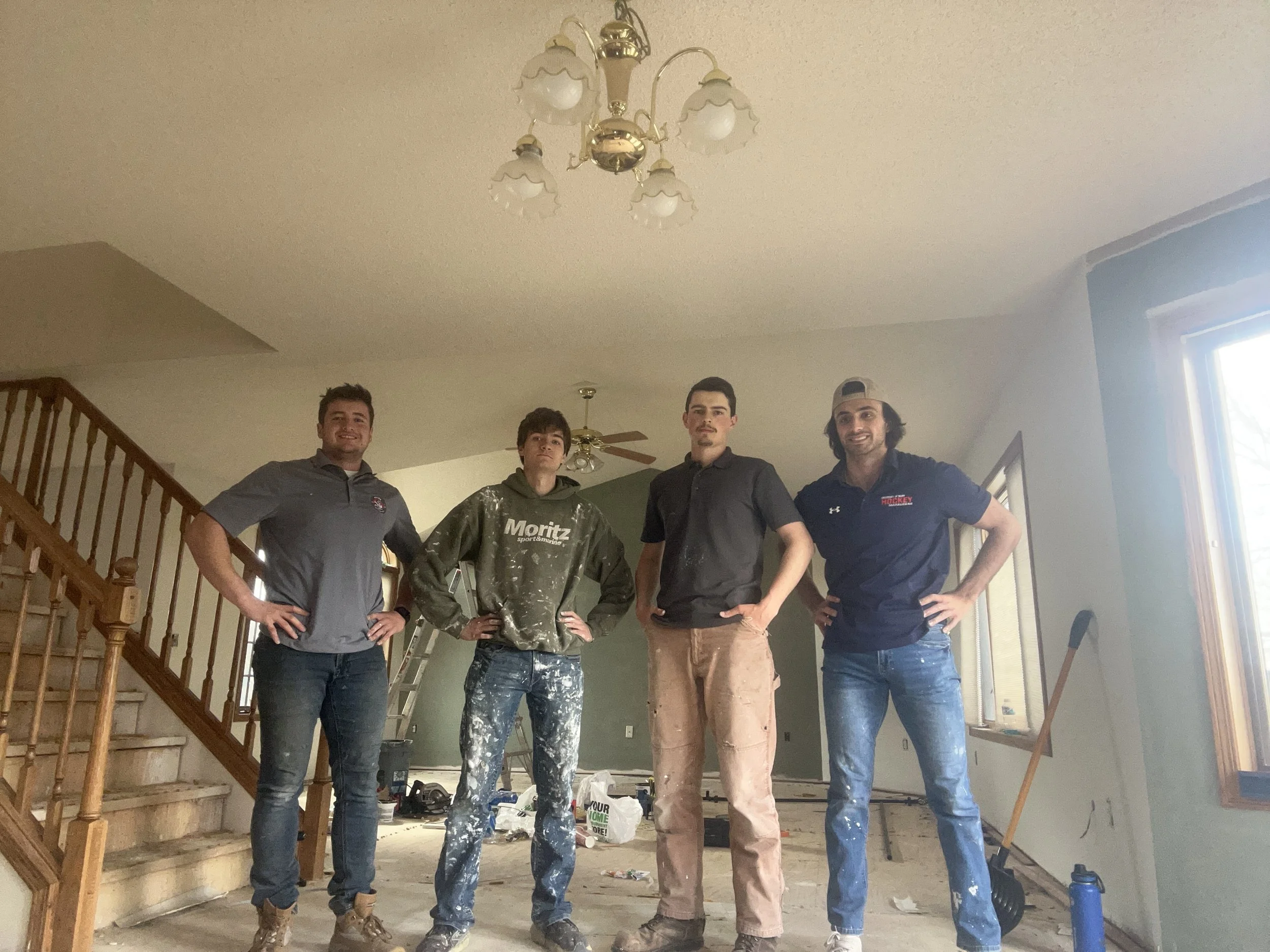 Four men standing inside a house under construction, posing for a photo. The area appears to be in the process of renovation or painting, with tools and supplies visible on the floor. A staircase is on the left, and there is a chandelier hanging from the ceiling.
