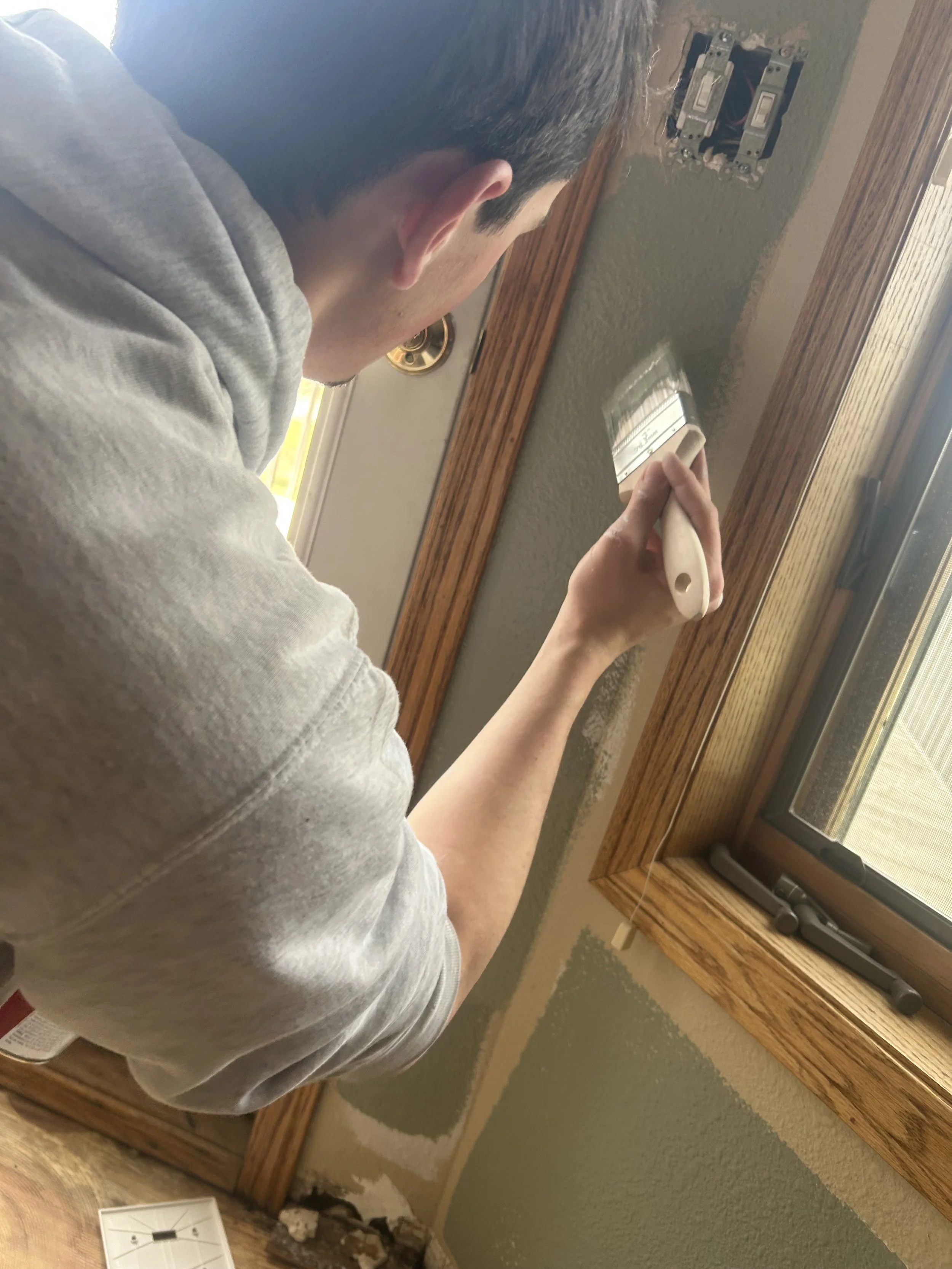 A person painting a wall near a window with a paintbrush, seen from behind and slightly to the side.