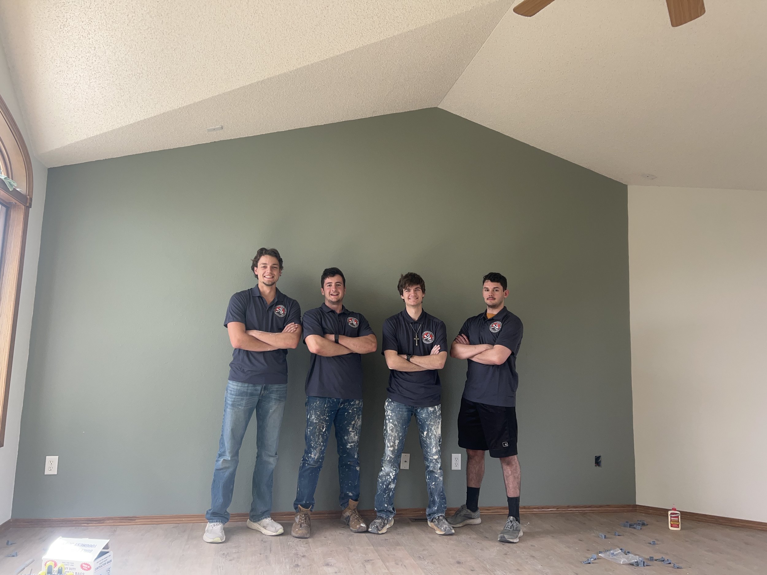 Four young men standing against a green wall in a room with large windows, wearing black shirts with a logo, and painted jeans or shorts, smiling and looking at the camera.