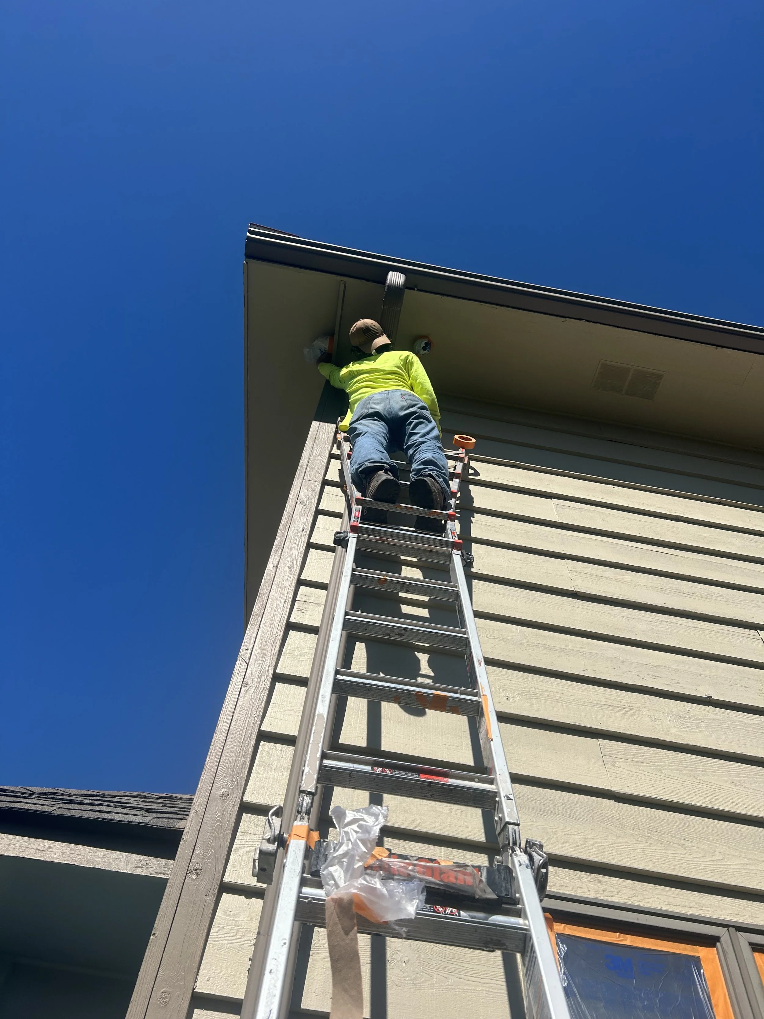 A worker in a yellow safety vest and hat standing on a ladder, working on the roof of a house against a clear blue sky.