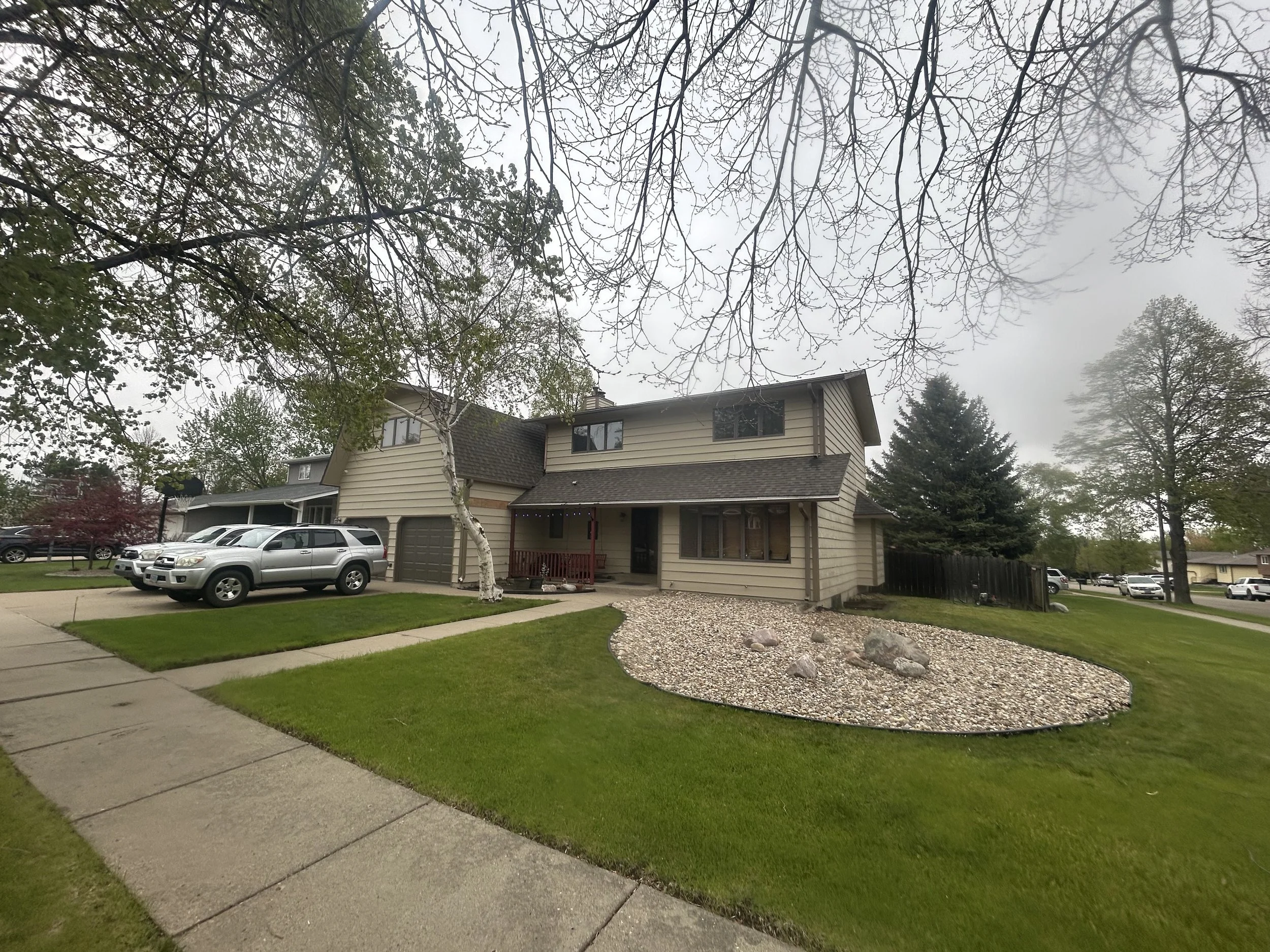 A suburban house with beige siding and dark roofing, with a front porch and multiple windows, surrounded by a neatly trimmed lawn and a curved garden bed with rocks. There are cars parked in the driveway. The scene is overcast with a cloudy sky and l