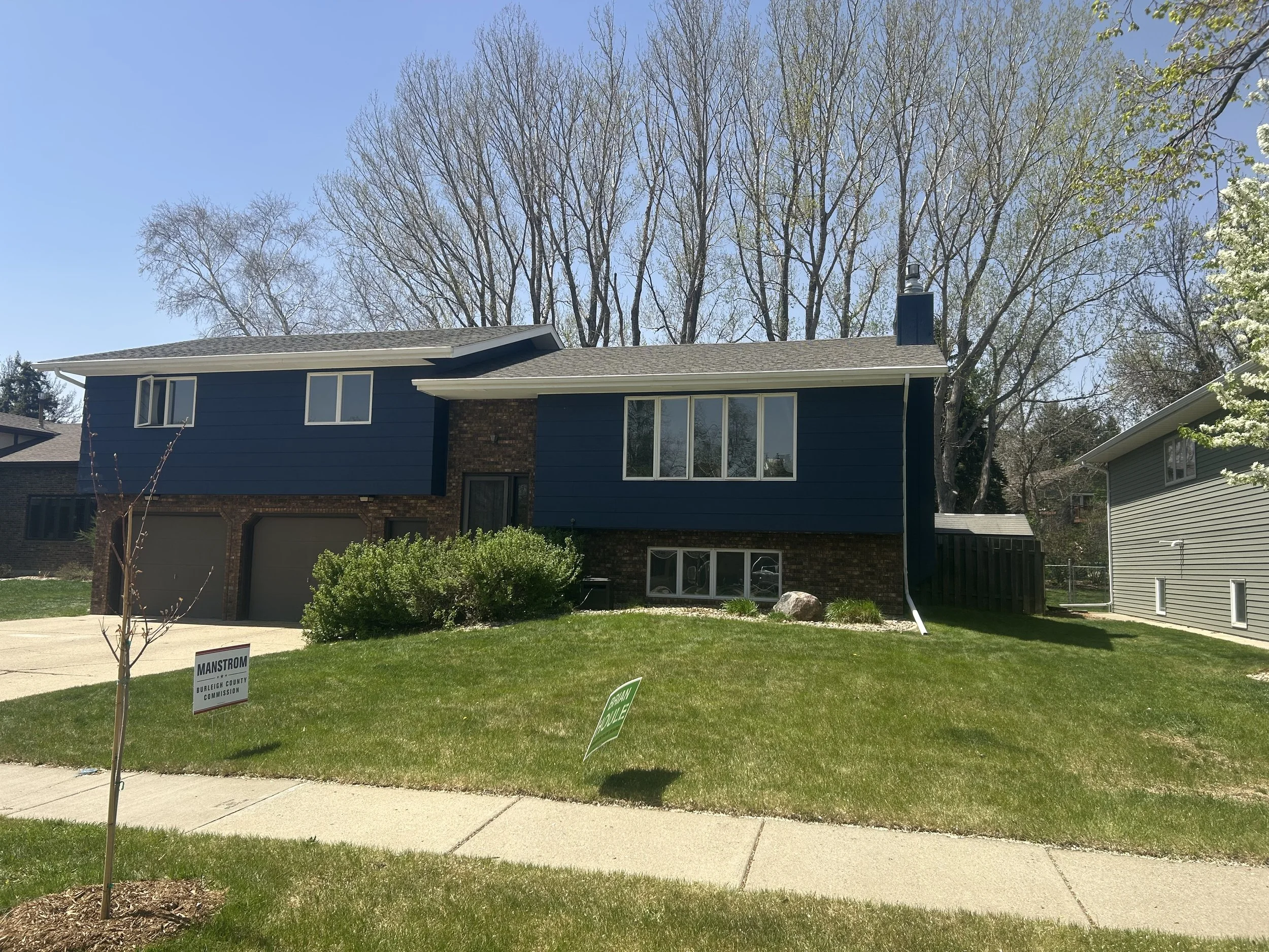A house with blue siding and a brick foundation, large windows, and a sloped roof, surrounded by green grass and trees in early spring. There are signs on the lawn indicating a local election or campaign.