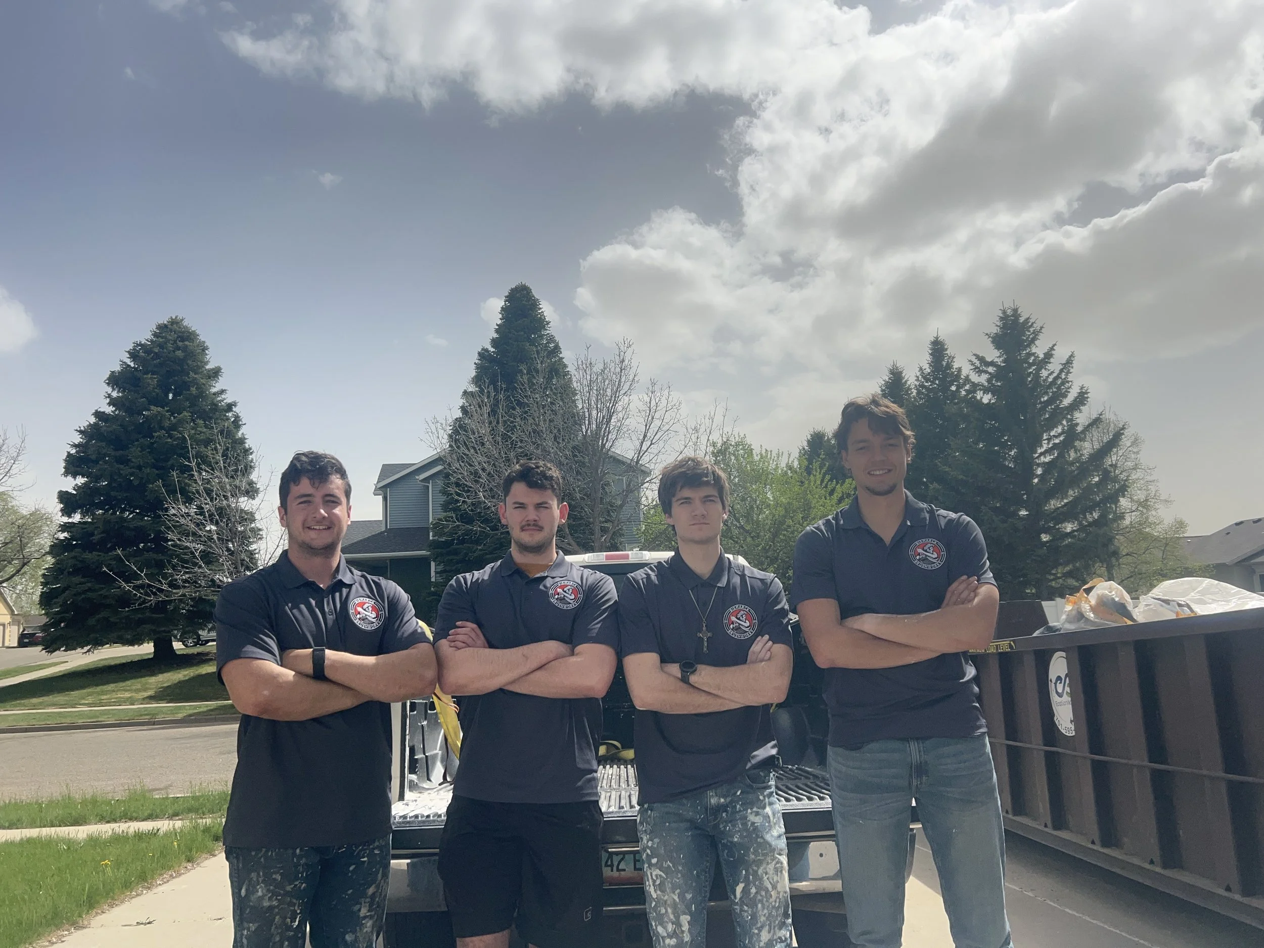 Four young men standing in front of a pickup truck outdoors on a sunny day, with trees and houses behind them, all wearing matching dark shirts with a logo on the chest.