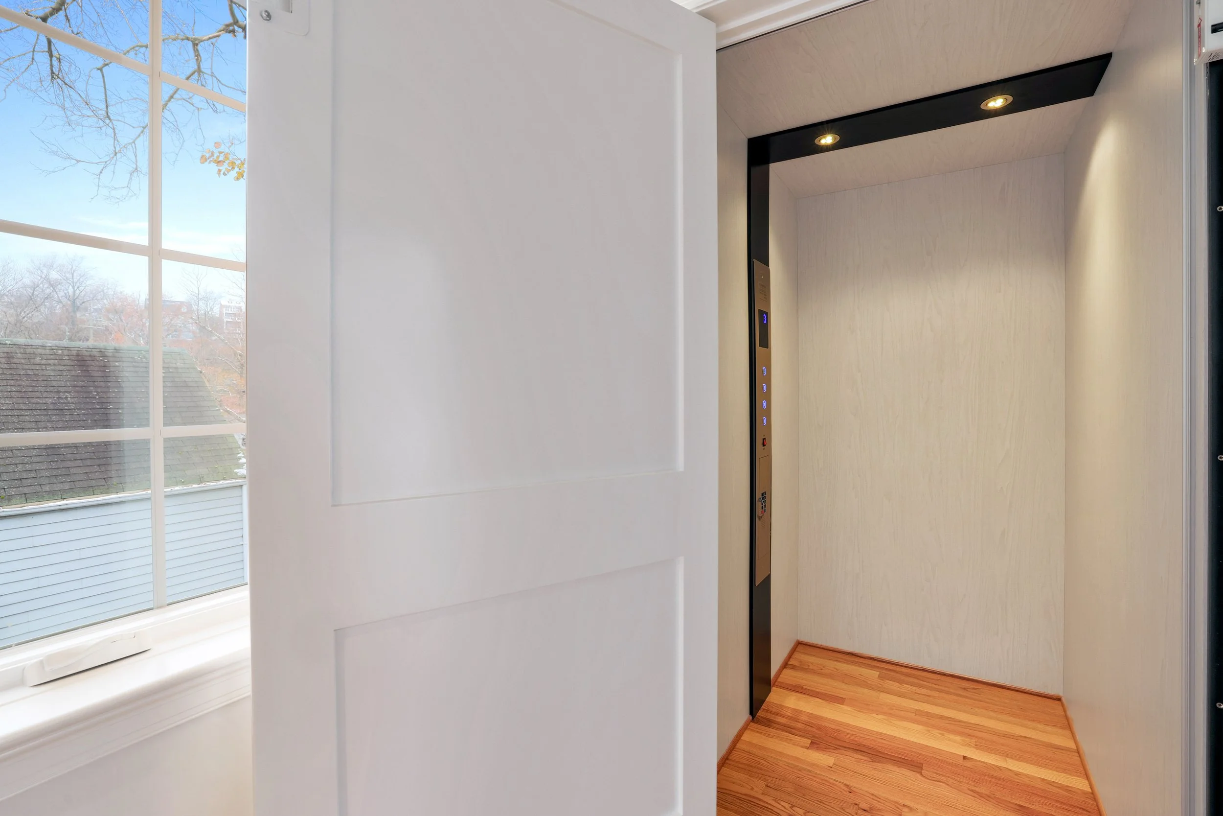 Interior view of an elevator with wooden flooring and beige walls, with a partially open door showing a window and an outside view of trees and rooftops.