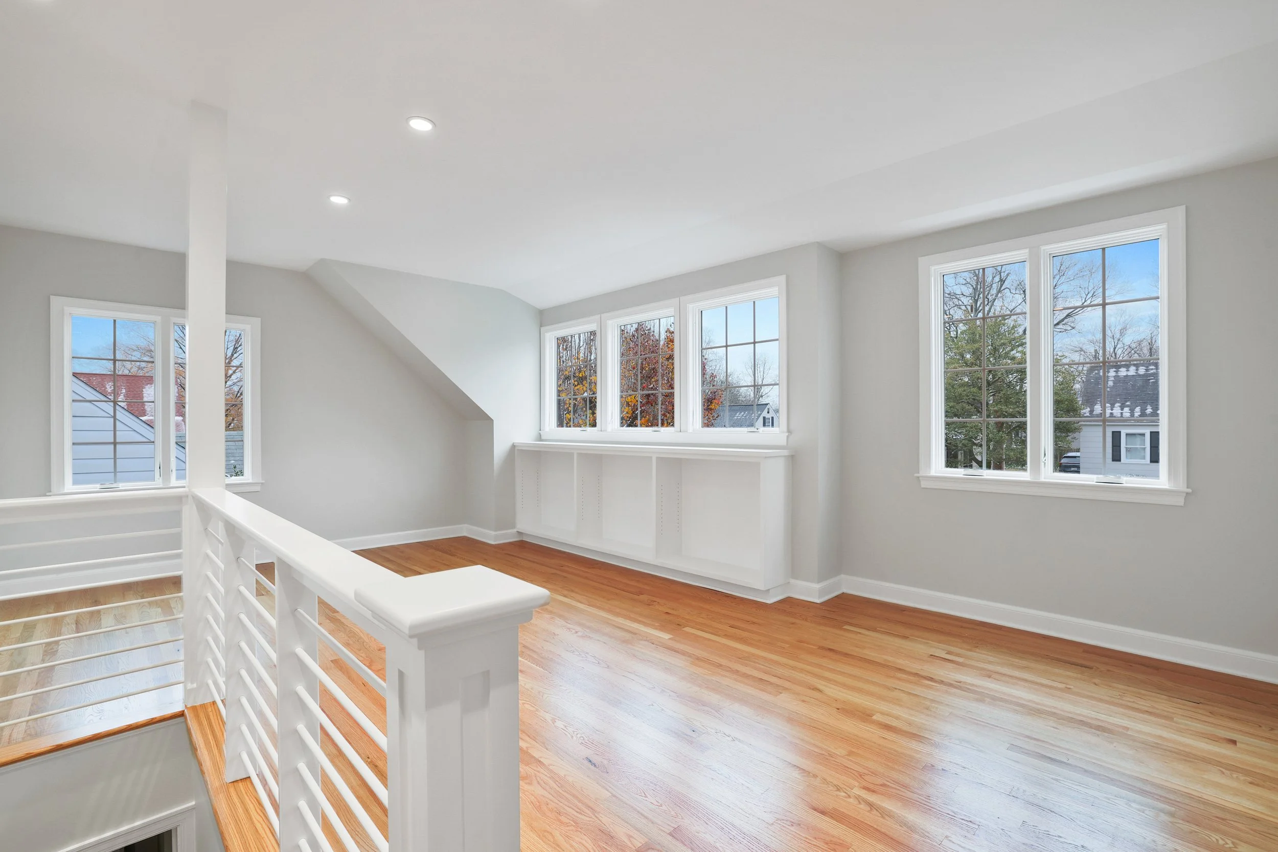 Bright, spacious empty room with hardwood floors, white walls, large windows, and a railing near the staircase. 