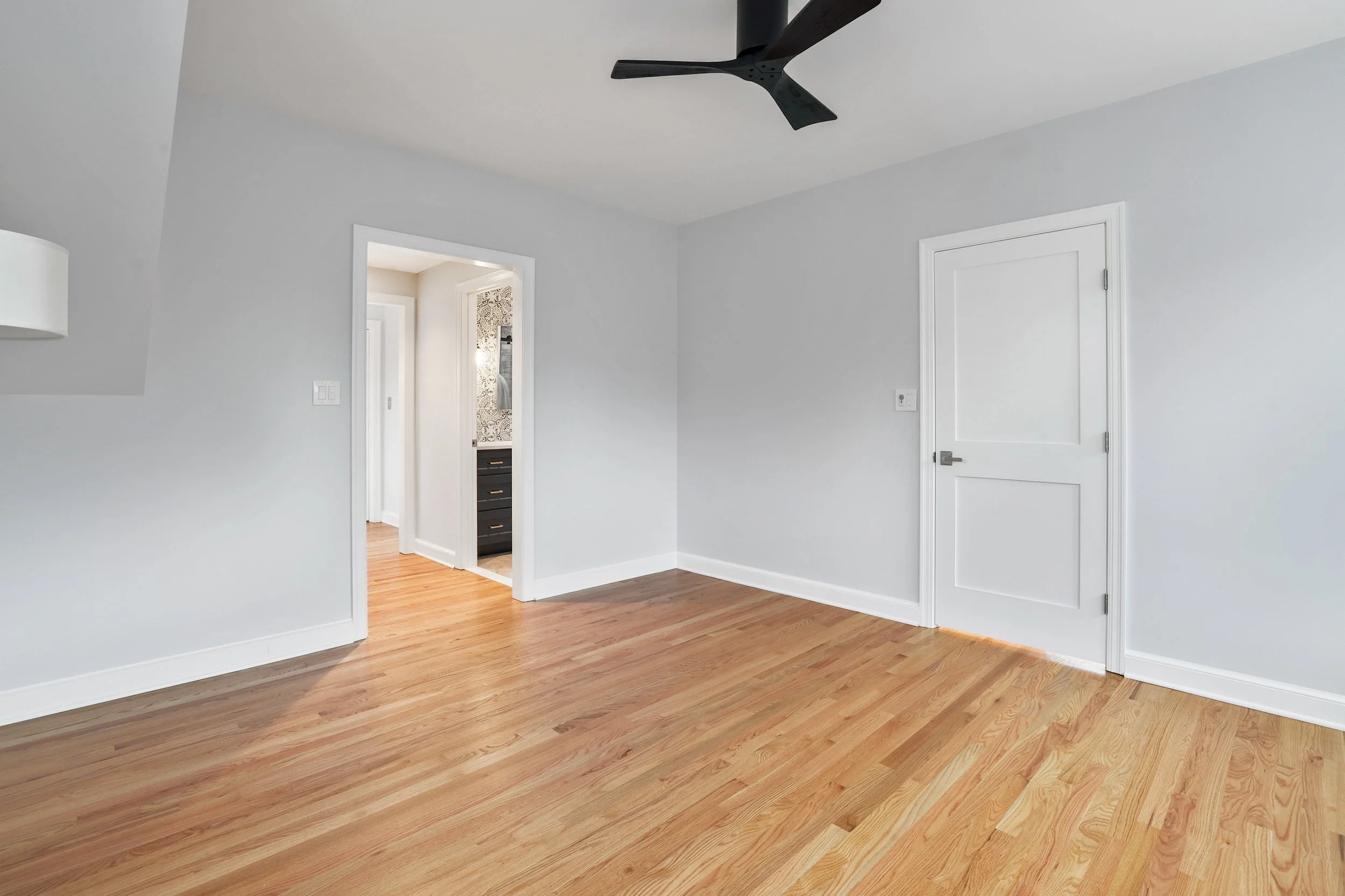Empty room with light gray walls, white trim, hardwood flooring, ceiling fan, and partially visible adjacent room with a black vanity and patterned wallpaper.