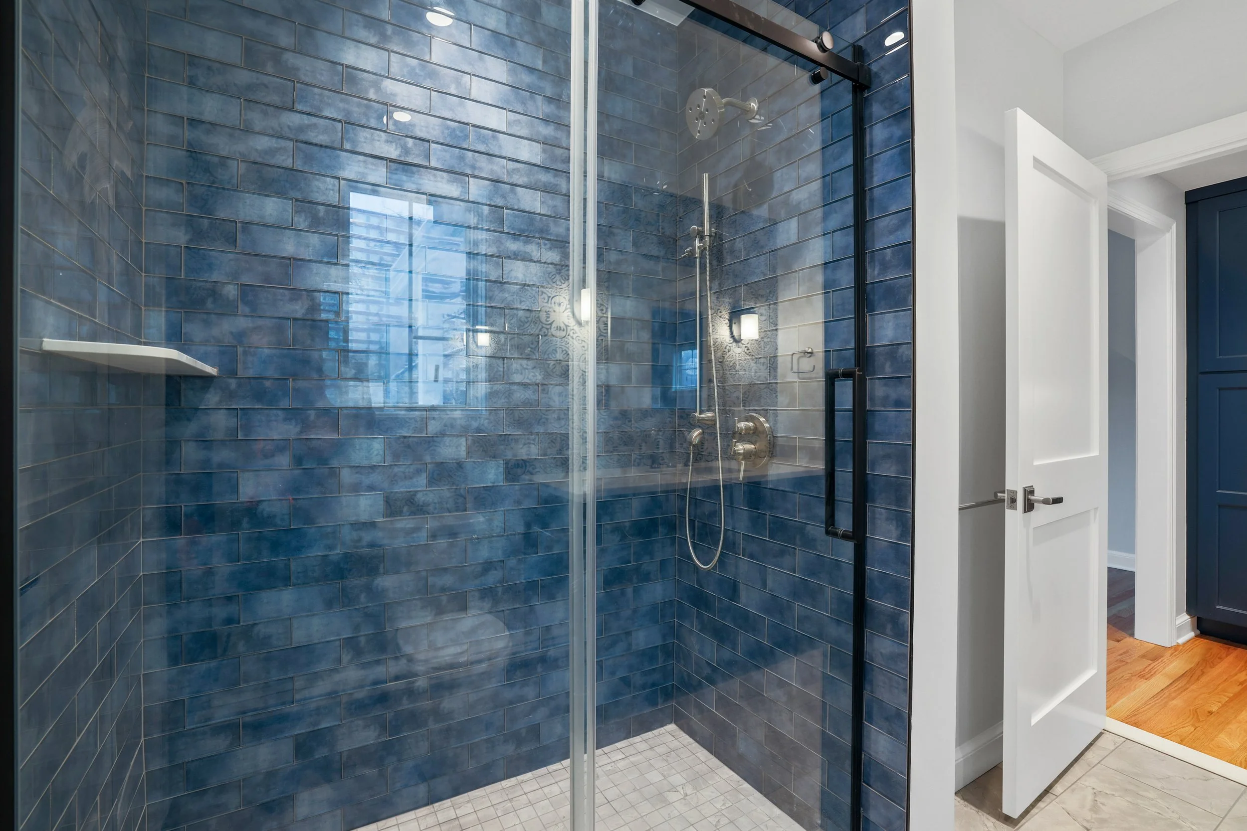 Blue tiled glass-enclosed shower with white and gray tiles on the floor, a showerhead, and a handheld shower. 
