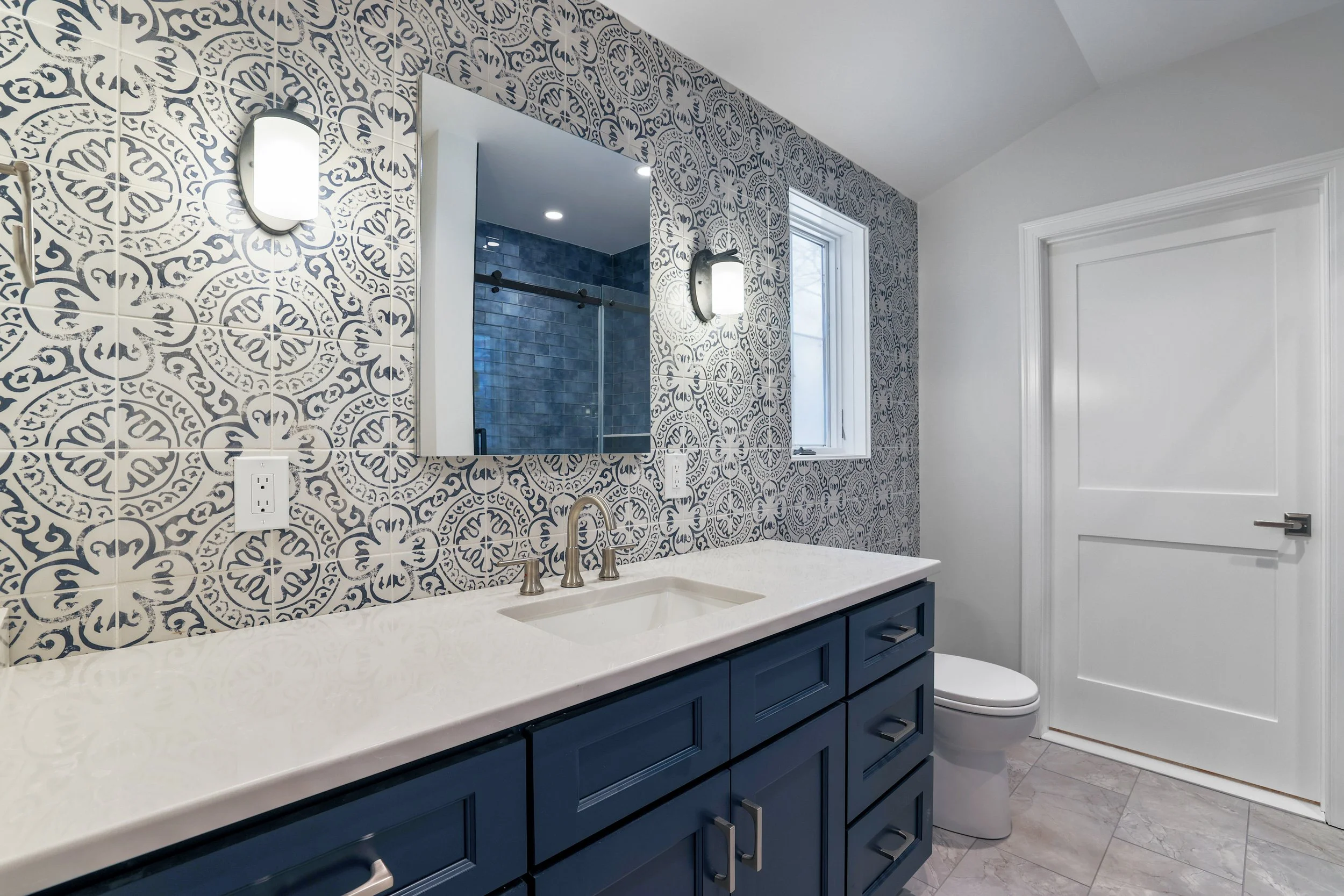 A bathroom with a navy blue vanity, a white countertop, a built-in sink, and brushed metal handles. The wall behind the vanity has patterned tile in black and white. There is a mirror above the sink, a window to the right, and wall-mounted lights on 
