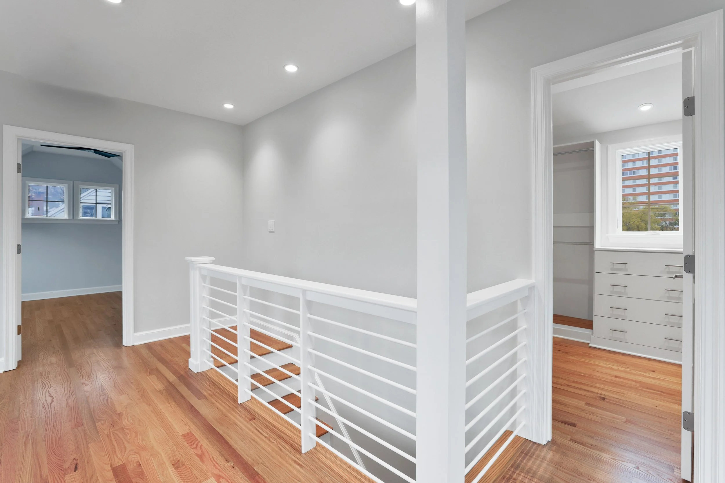 Interior view of a modern home with white walls, wooden floors, and a white railing around a staircase, including doorways leading to other rooms.
