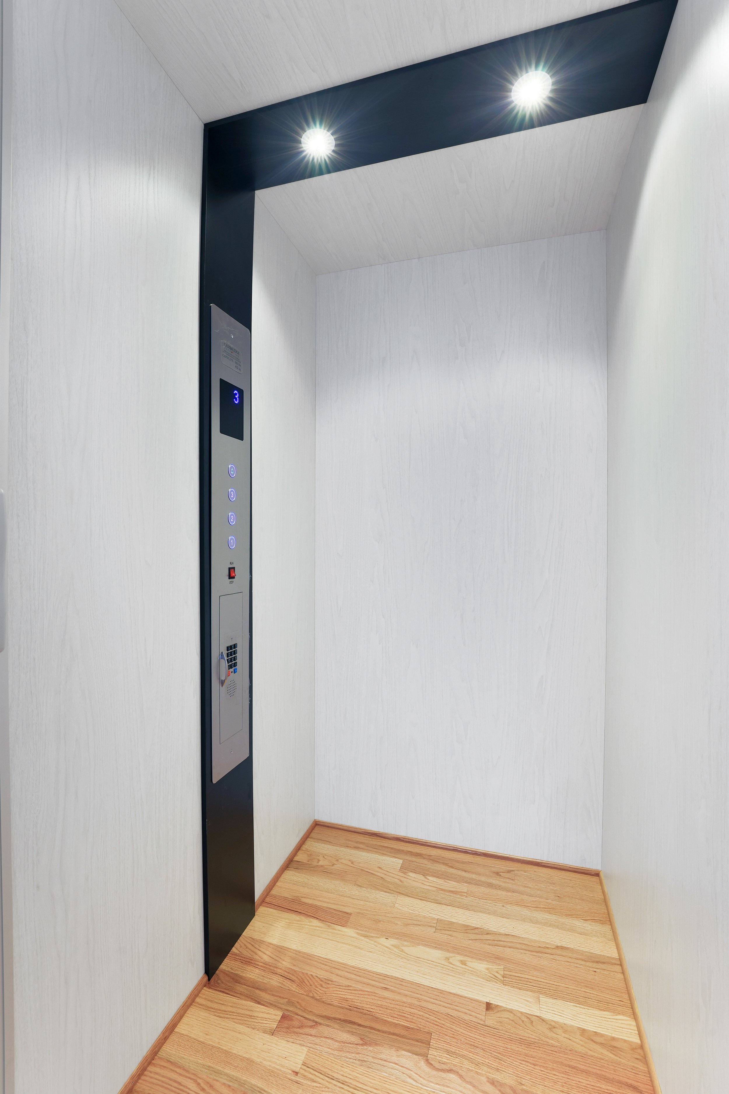 Empty elevator with wood flooring and white walls, illuminated by two bright ceiling lights.