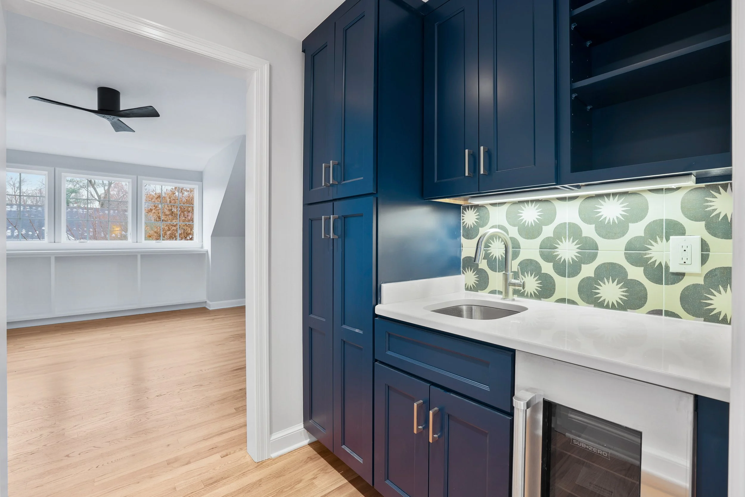 Master bedroom coffee bar with blue cabinets, a small sink, patterned tile backsplash, and a bit of the adjoining room with wooden flooring and large windows.
