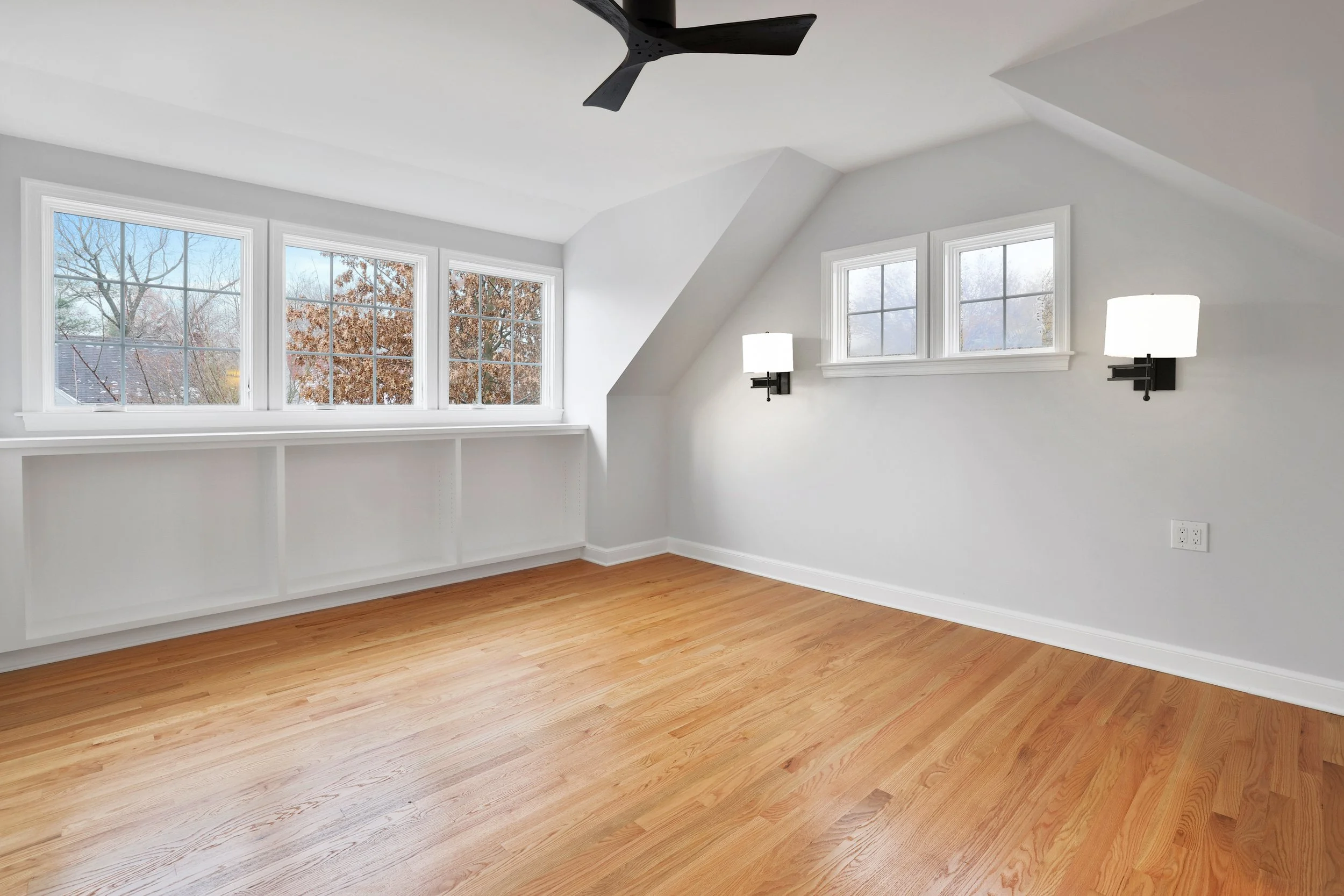 An empty room with white walls, hardwood floors, multiple windows, wall-mounted lamps, and a ceiling fan.