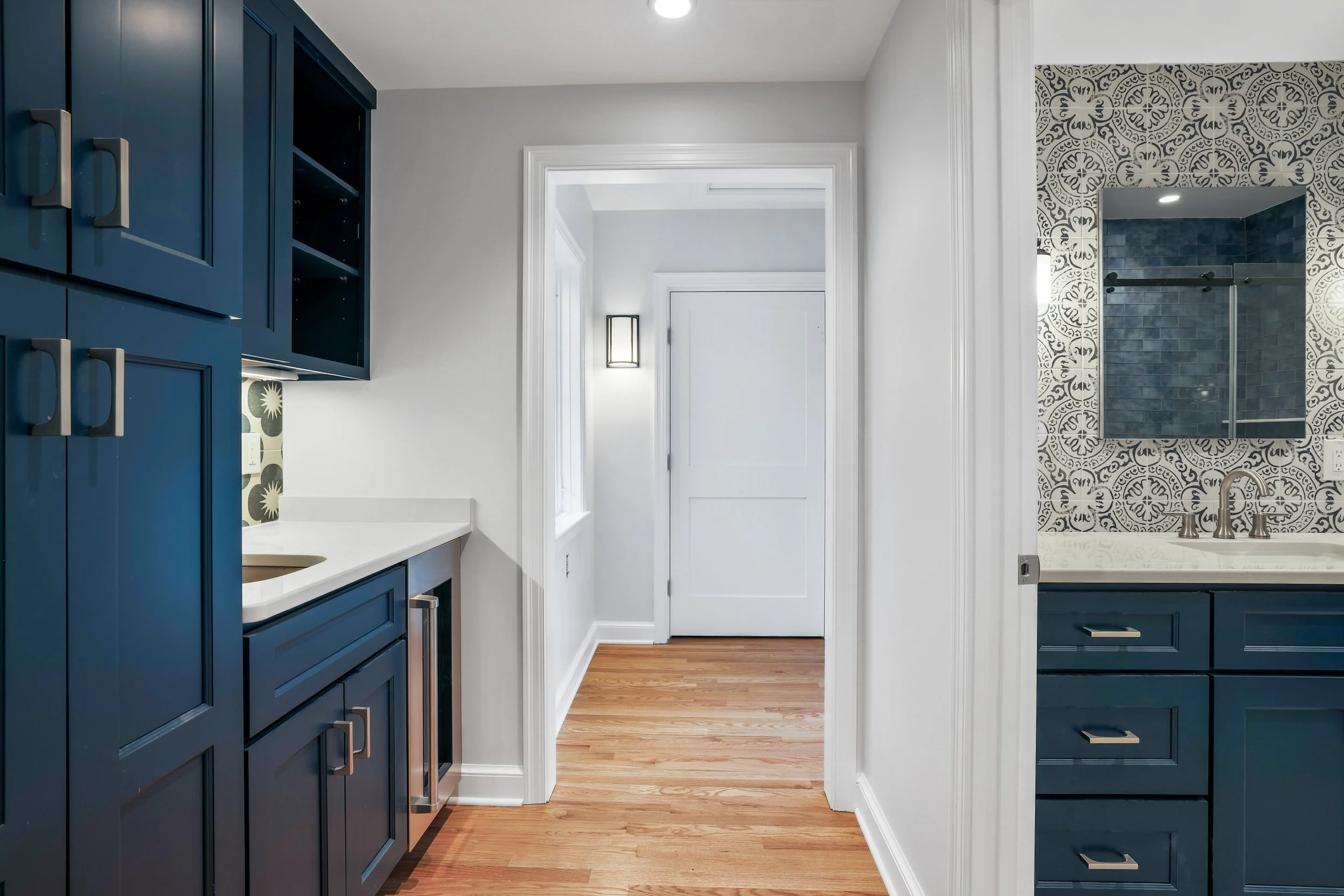 Interior view of a modern kitchen with dark blue cabinets, white countertops, and a hallway with white walls and hardwood floors.
