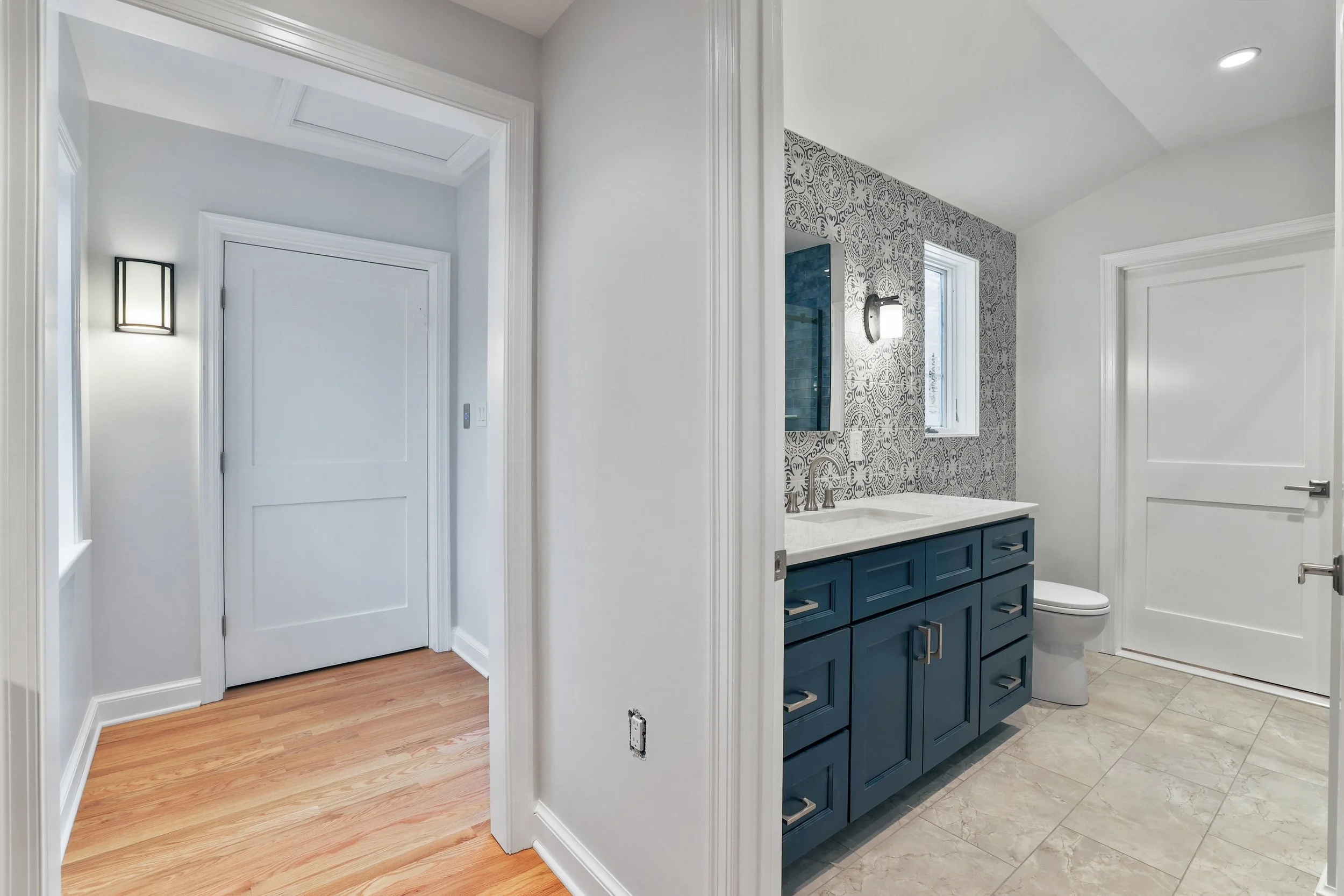 View of a bathroom with a blue vanity, patterned backsplash, and a window, adjacent to a hallway with a closed white door with elevator hidden behind and light hardwood flooring.