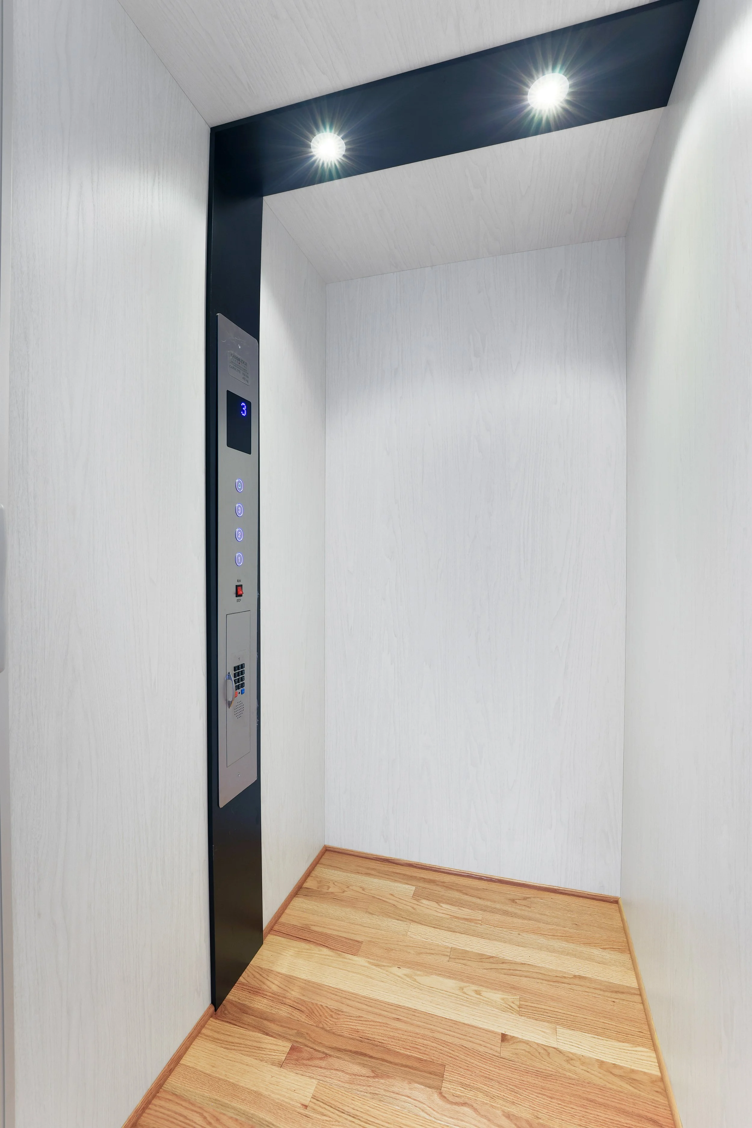 Empty elevator interior with two ceiling lights, control panel, and wooden flooring.