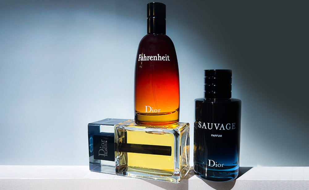 Three Dior perfume bottles on a white surface with a plain blurred background. The bottles are labeled Fahrenheit, Sauvage, and another Dior perfume, with the Sauvage bottle being black and blue, and the Fahrenheit bottle having a gradient from red to yellow.