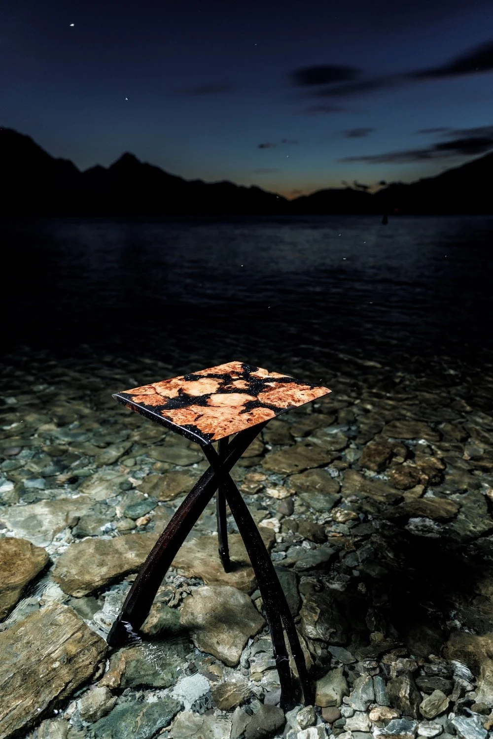A small, decorative table with a marbled top on a rocky shore at dusk, with mountains and a dark sky with stars in the background.