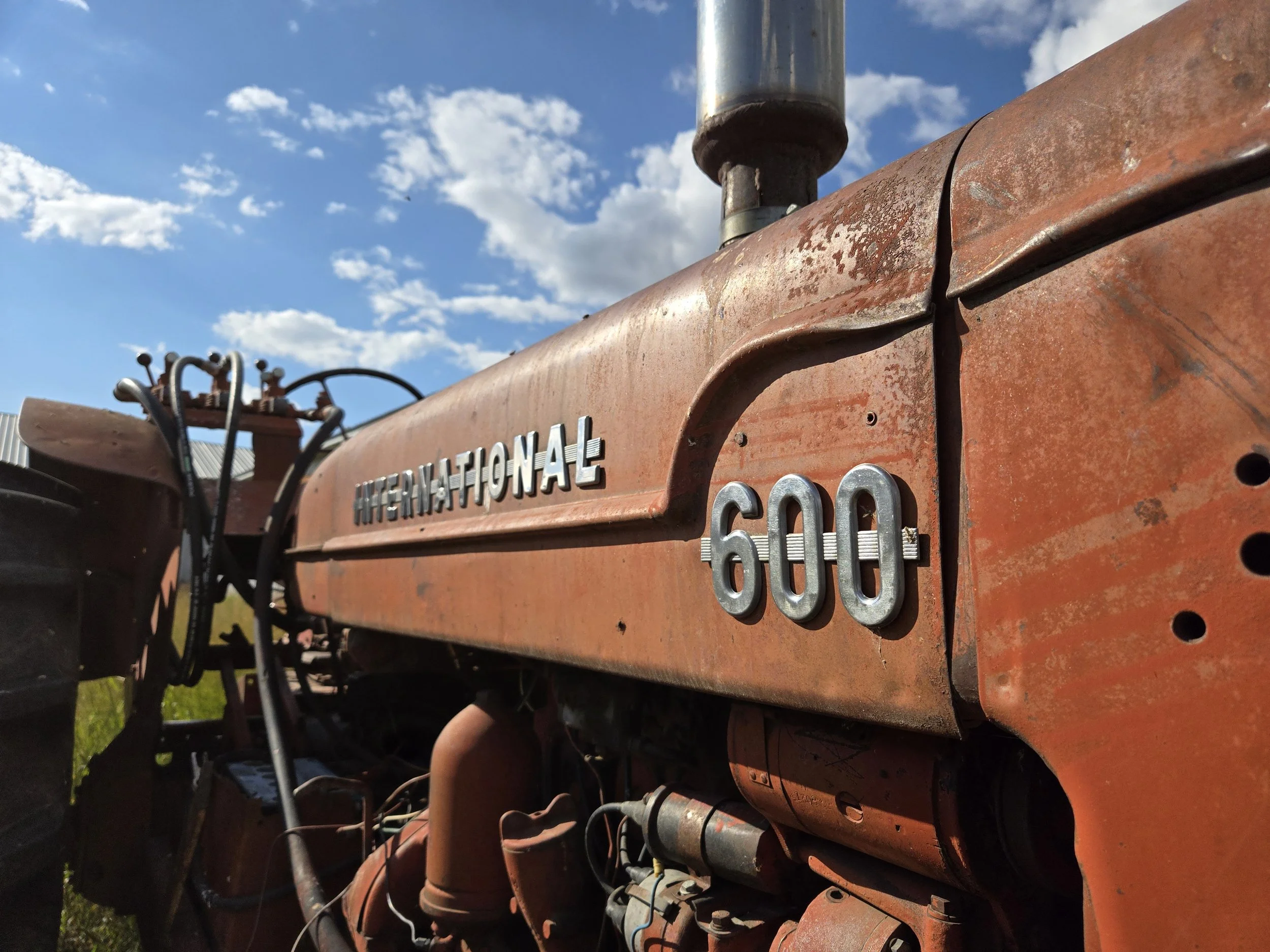 Close-up of an orange International 600 tractor with rusted metal, showing the side with the brand name and model number, against a blue sky with clouds.