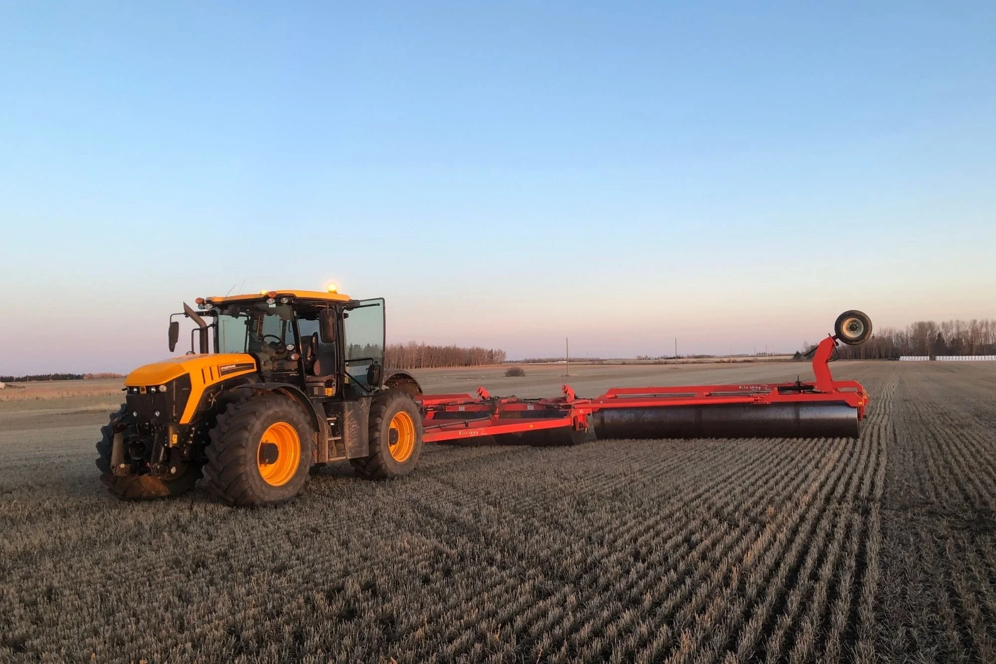 A JCB fastrac 4220 pulling a land rollert working in a large field during sunset.