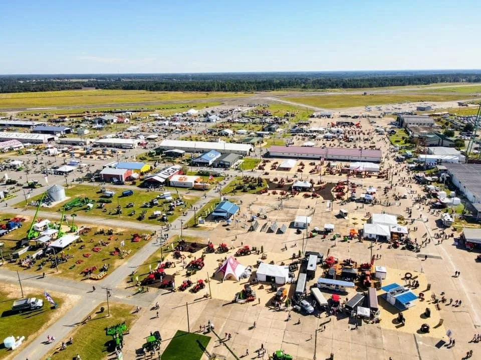 Aerial view of a large outdoor event or fair with tents, booths, and vintage tractors on a concrete and grassy area, with open fields and a runway in the background.
