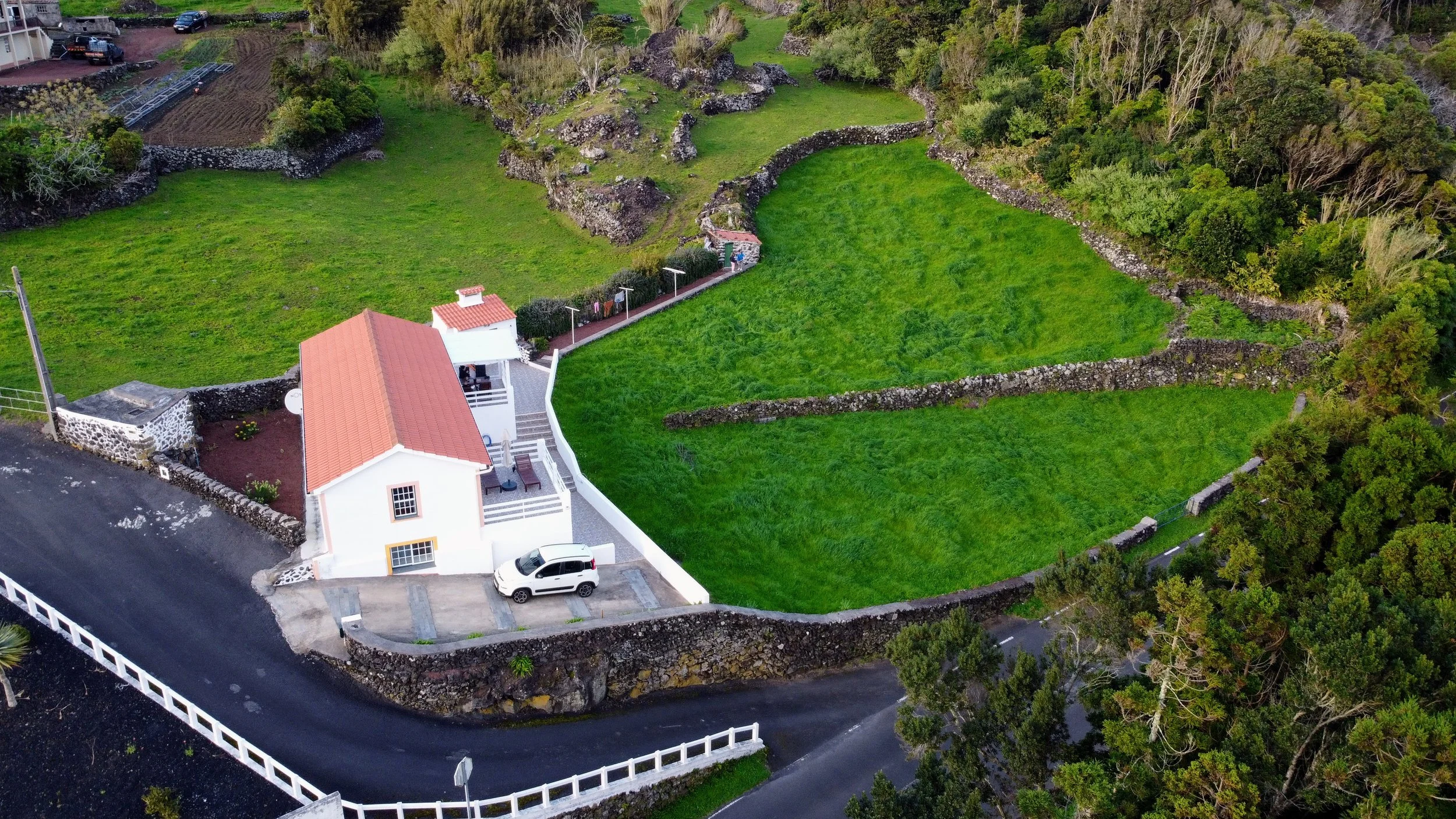Aerial view of a white house with a red tile roof situated in a lush green landscape, featuring a manicured lawn, stone walls, trees, and a winding paved road.