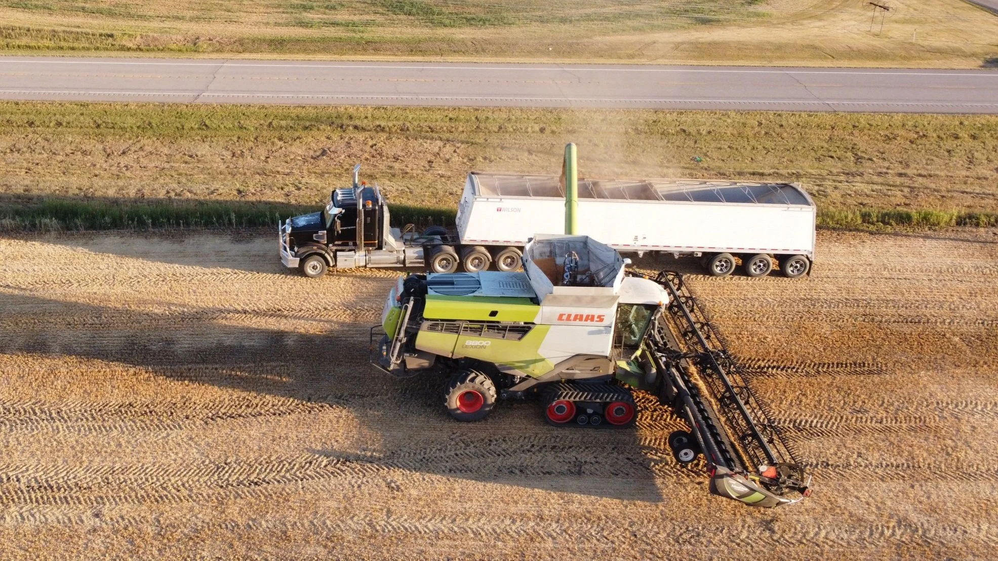 An aerial view of agricultural machinery working in a field with a road in the background. There is a large CLAAS combine harvester and a semi-truck with a trailer, harvesting and transporting crops.
