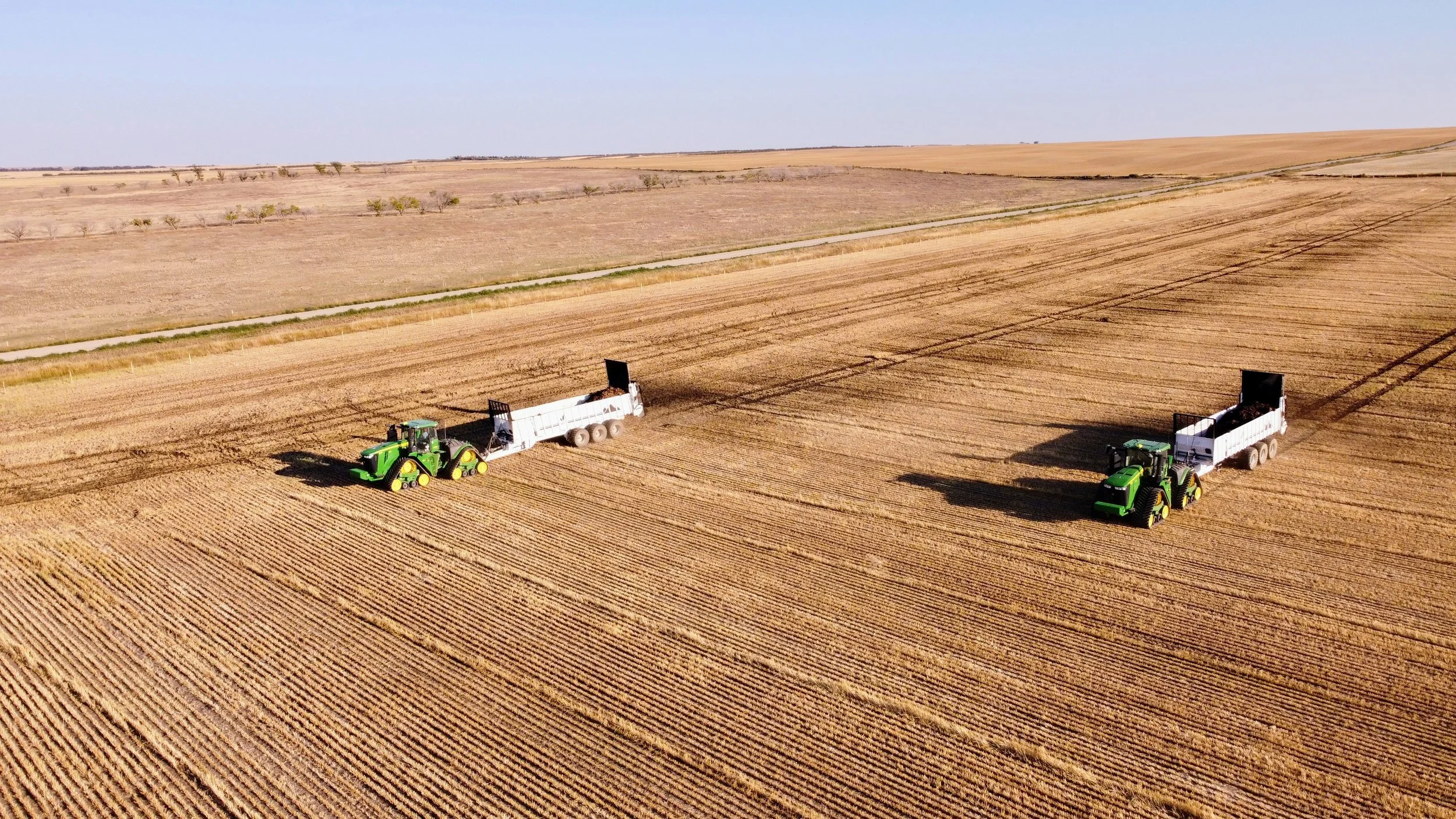 Two green tractors harvesting crops in a large, open field with clear skies.