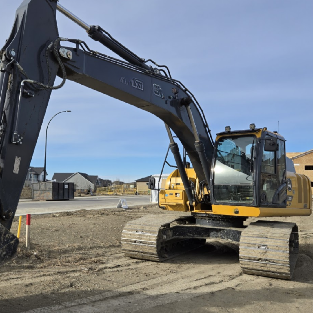 A yellow John Deere excavator on a construction site, with residential houses and a clear blue sky in the background.